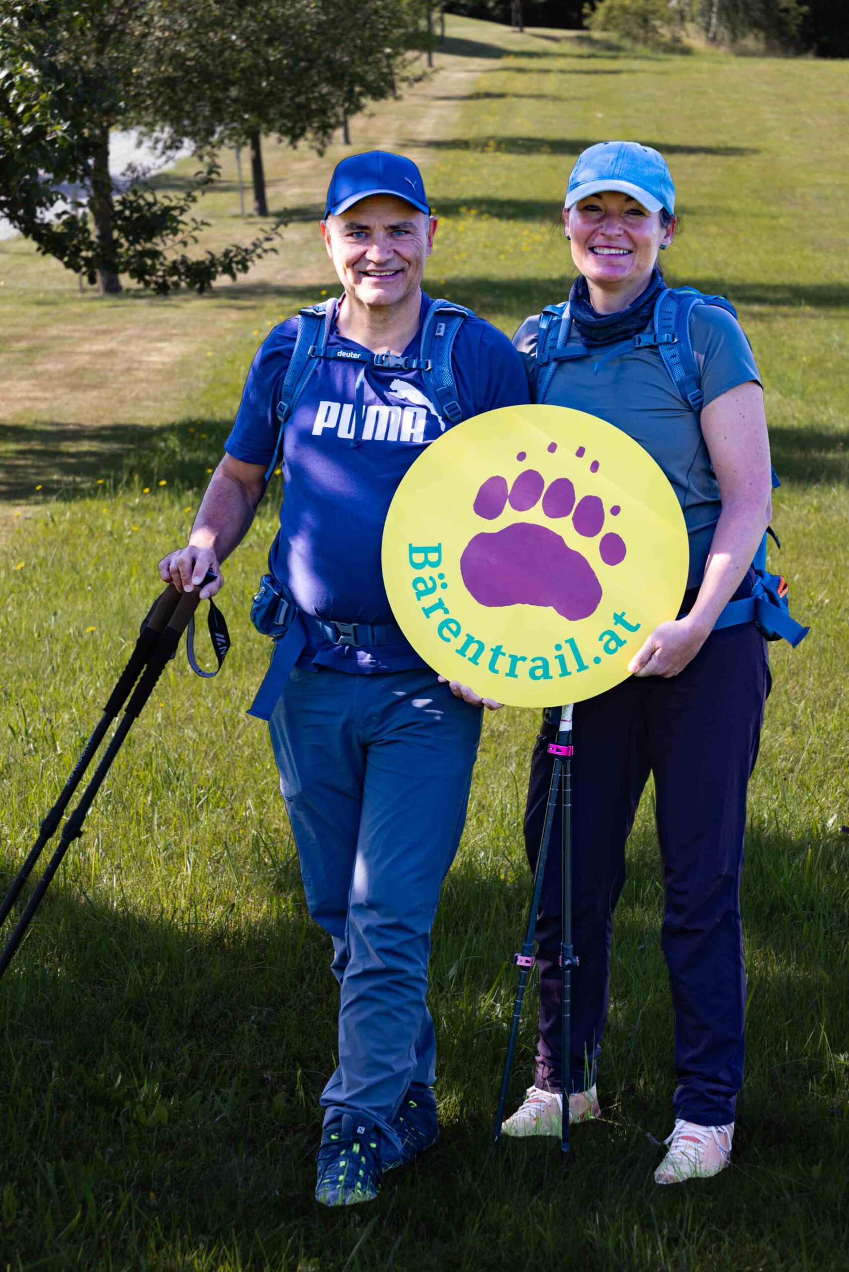 Wanderpaar in voller Ausrüstung mit Bärentrail-Logo auf einer Wiese