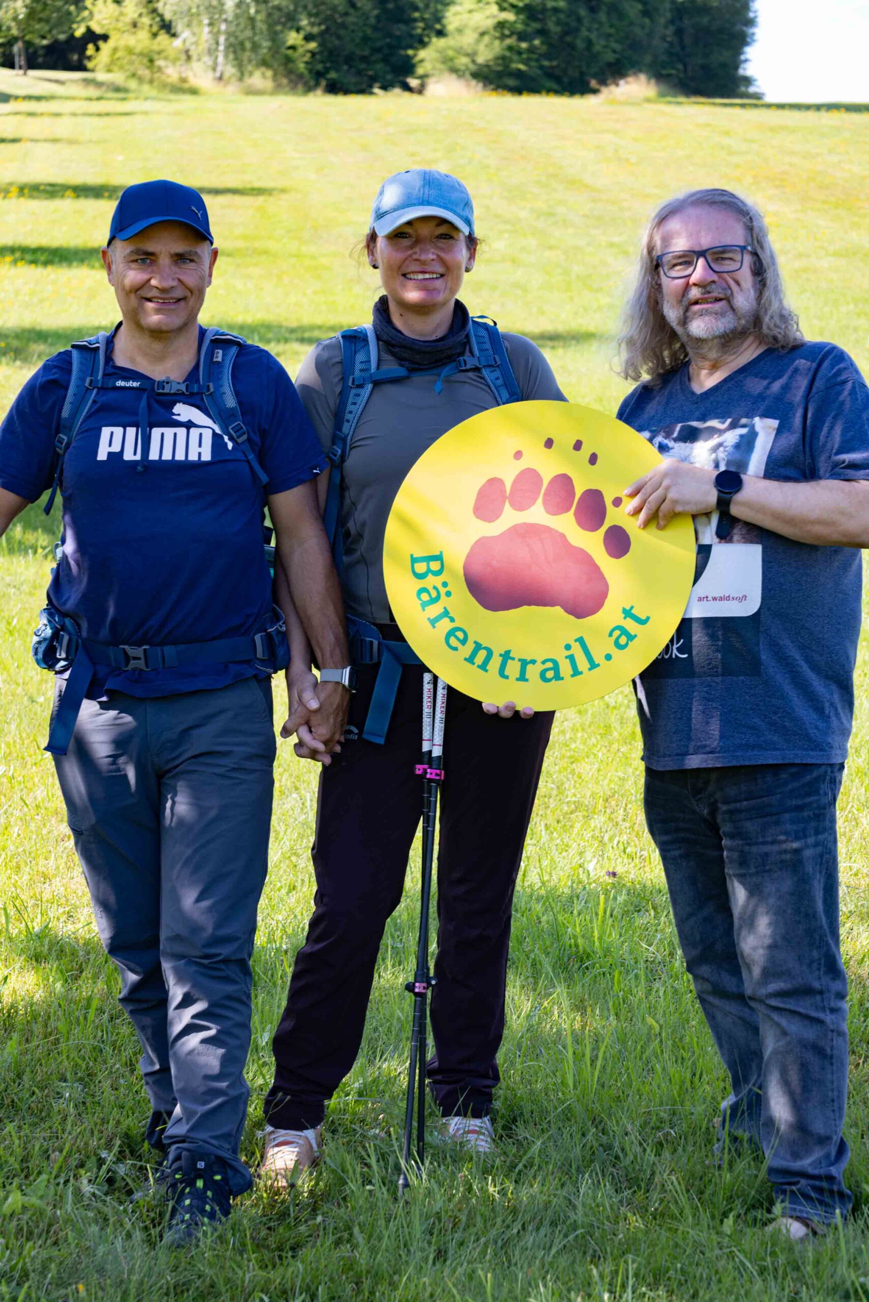 Zwei Wanderer und Gastgeber Martin Lugmayr halten gemeinsam das Bärentrail-Schild auf einer Wiese
